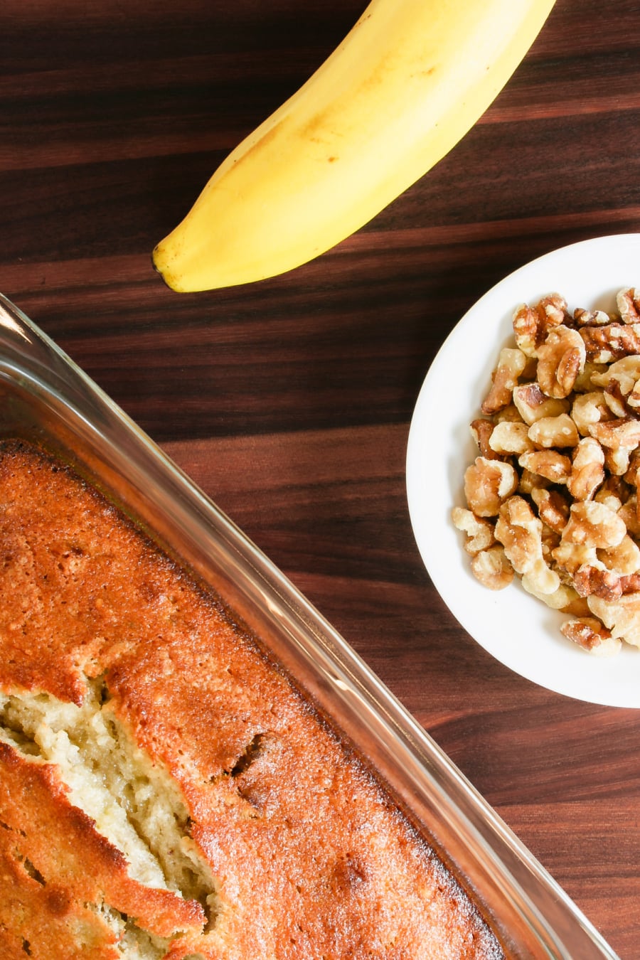 banana nut bread in a baking dish next to a banana and a bowl of walnuts