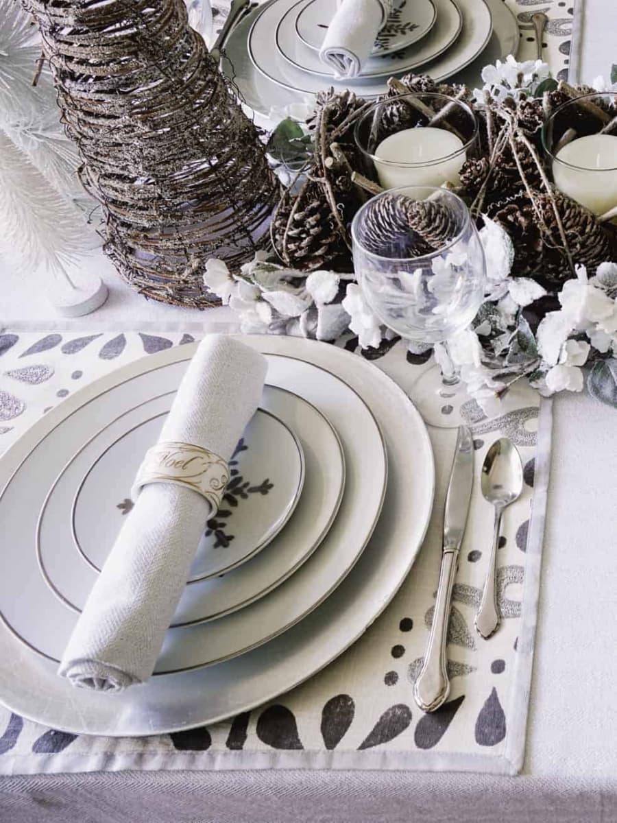 A Christmas place setting in the dining room featuring a charger, snowflake designed plates, a napkin, and Christmas napkin ring, sitting on a placemat with silver accents in the design. In the background a table centerpiece is setup with candles and surrounded with frosted florals.