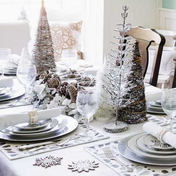 Dining room table Christmas decor featuring silver placemat, silver charger, and plates with silver snowflake pattern, white, sparkly napkins with Christmas napkin rings, decorative snowflake coasters and a centerpiece with pine cones and white candles flanked by a number of decorative trees.