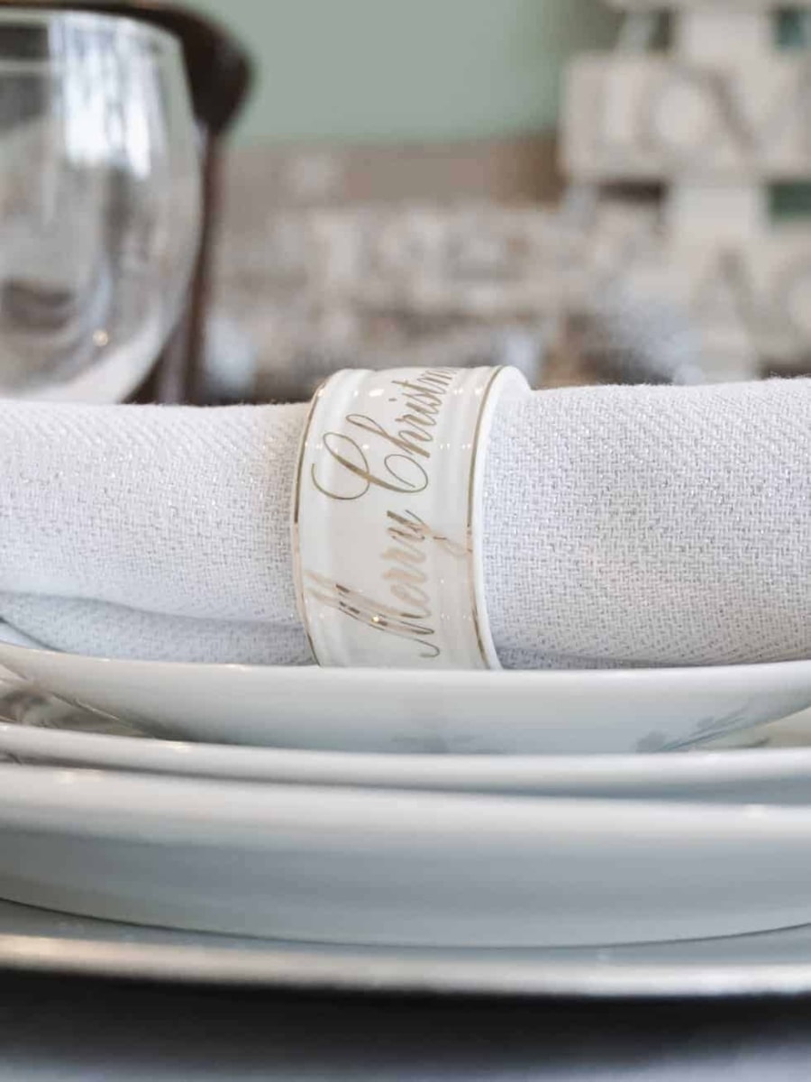 A Merry Christmas napkin ring holding a white napkin and sitting on top of a stack of Christmas plates.