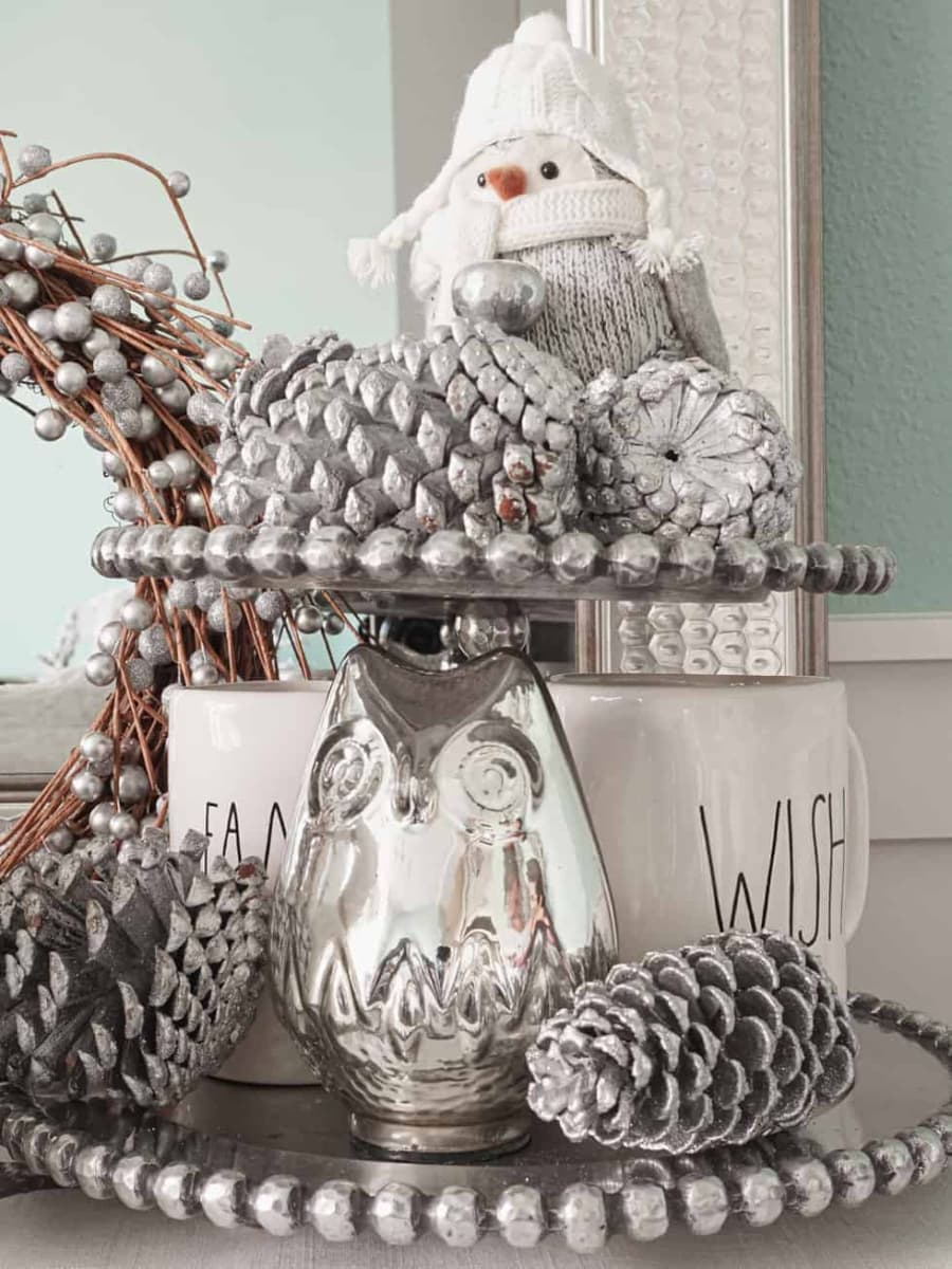 A silver tiered tray with silver pine cones, Rae Dunn mugs, and cloth bird, and silver owl.