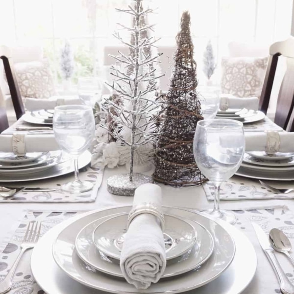 A beautiful dining room Christmas place setting with a stack of Christmas plates, napkin and Christmas napkin holder. A table centerpiece with decorative trees and candles can be seen in the background.