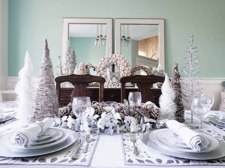 A Christmas dining room featuring a fully set table with pine cone and candle centerpiece, Christmas plates, placemats, and napkins. The buffet is also fully decorated in Christmas wreaths, decorative Christmas trees, and Christmas animals.