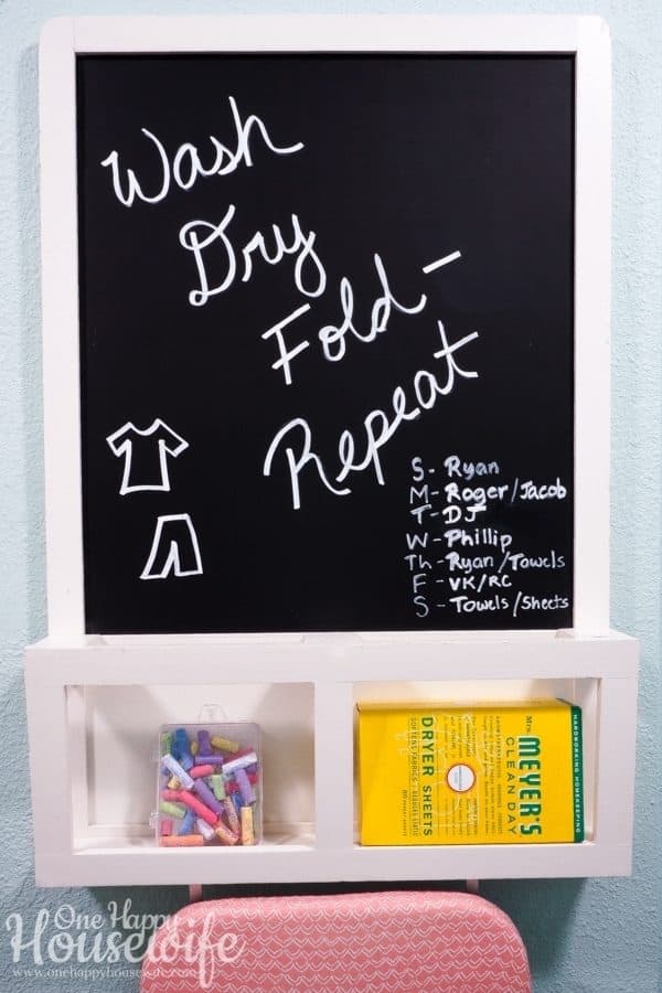 A chalkboard hanging in the laundry room with the words Wash, Dry, Fold, Repeat written on it along with a daily laundry schedule.