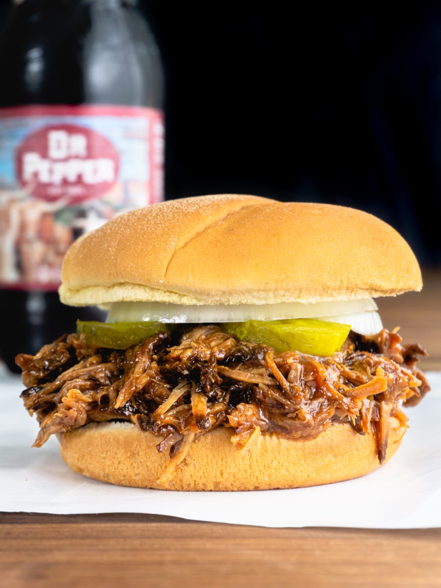 a dr pepper pulled pork topped with pickle and onion slices served with a bottle of dr pepper