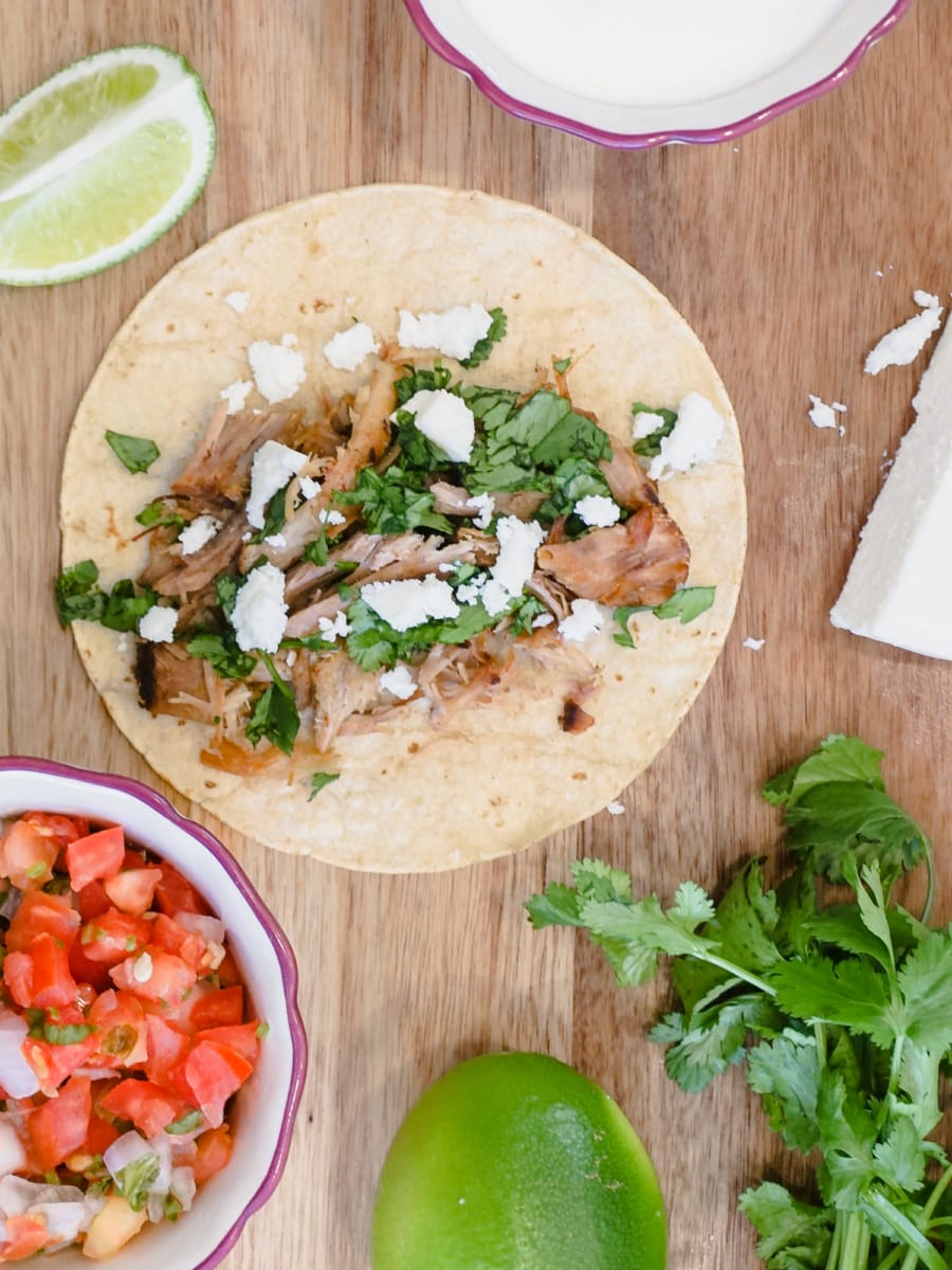 a corn tortilla with carnitas cilantro and crumbled cheese sitting on a cutting board with a bowl of pico de gallo, a lime slice, and a bowl of sour cream