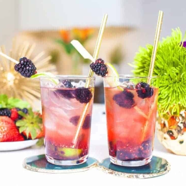 a non-alcoholic blackberry mojito served in two different cups next to a plate of different berries