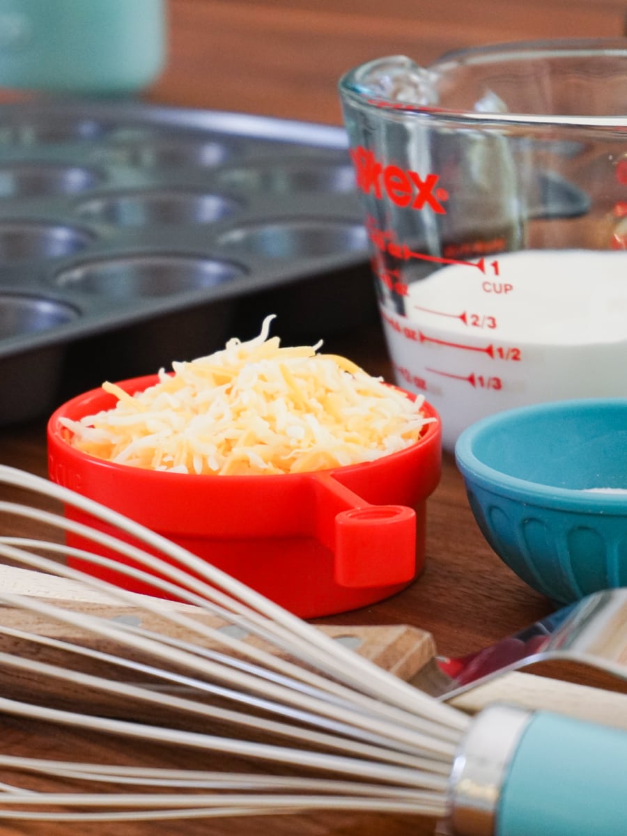 a mini muffin tin next to a whisk and shredded cheese