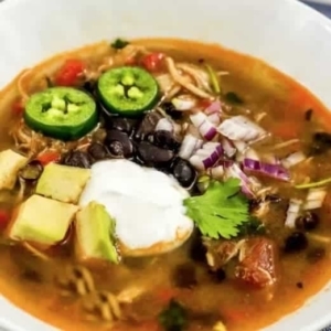 close up of a bowl of chicken fajita soup topped with sour cream, avocado slices, and two jalapeno slices