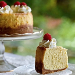 a white chocolate raspberry swirl cheesecake on a cake stand next to a plate with a slice of cheesecake