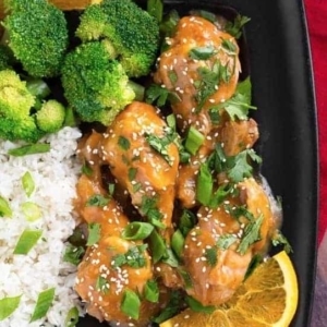 orange chicken legs garnished with scallions, cilantro, and sesame seeds on a plate with broccoli and rice