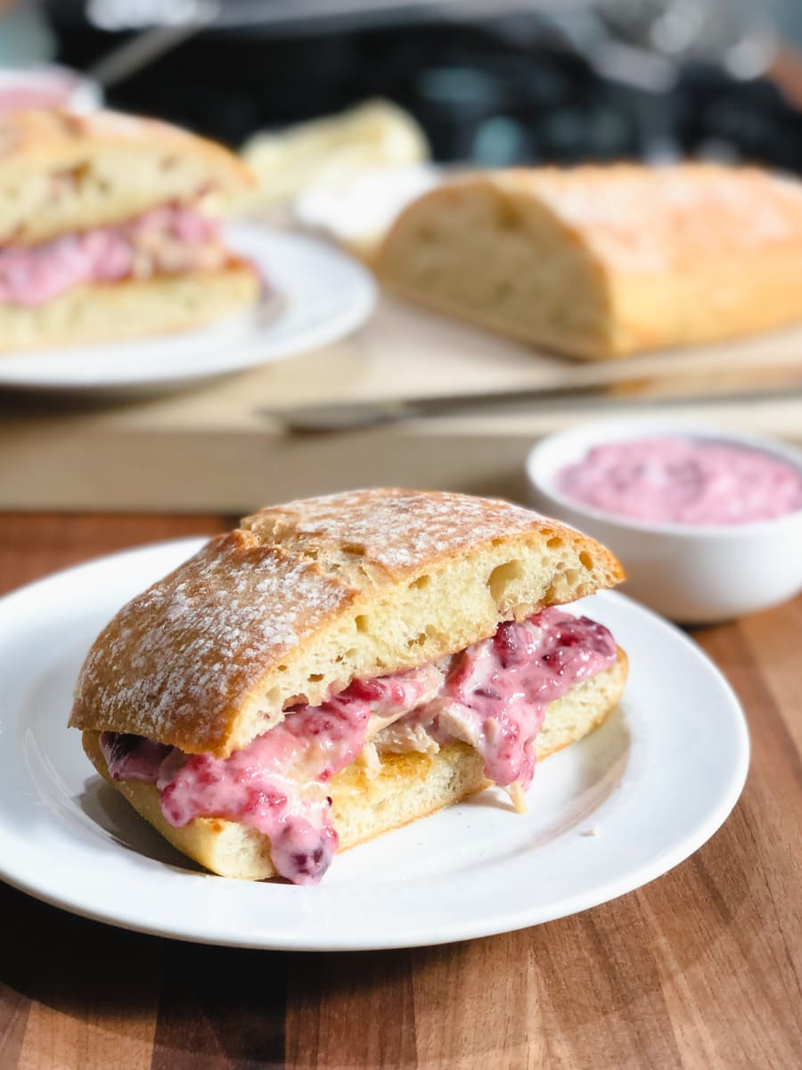 a leftover turkey sandwich with cranberry aioli sitting on a plate with a loaf of bread and small bowl of cranberry aioli in the background