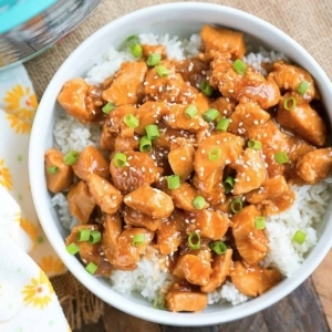 a bowl of sesame chicken on top of rice garnished with scallions