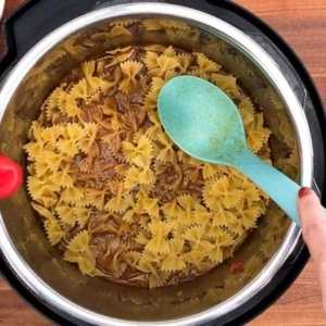 gently pushing the bowtie pasta down to submerge it into the beef broth using an aqua silicone spatula