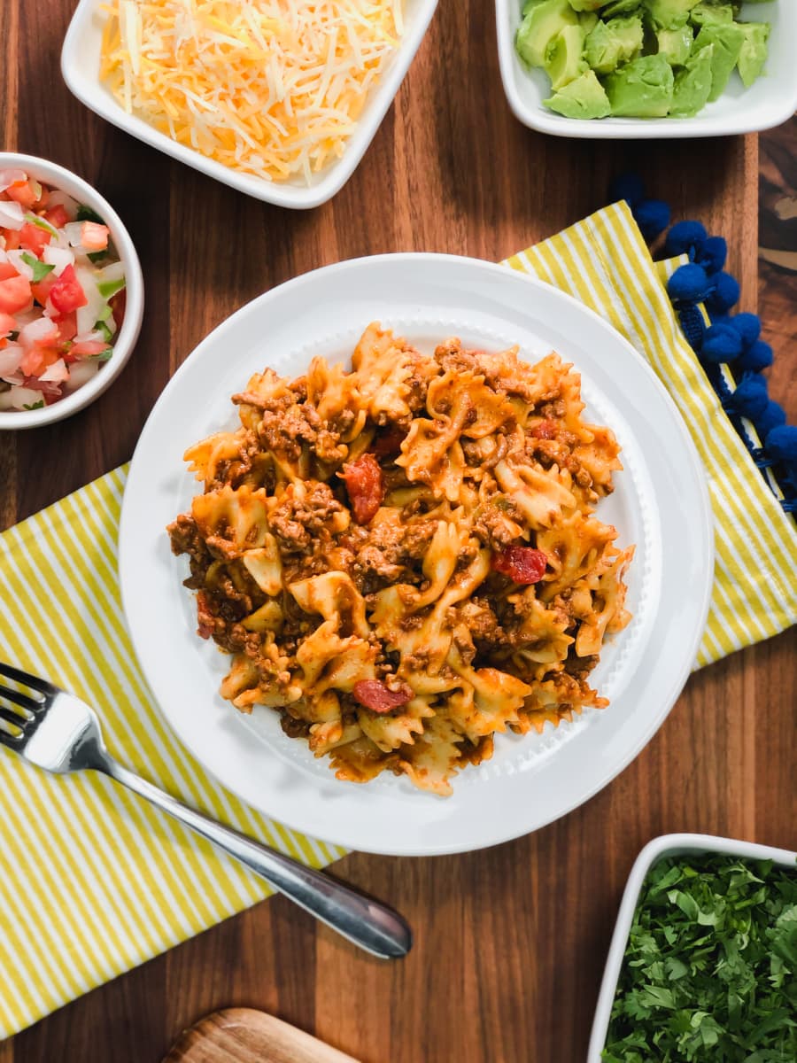 a plate of taco pasta along with small bowls of toppings including avocado, pico de gallo, shredded cheese, and cilantro