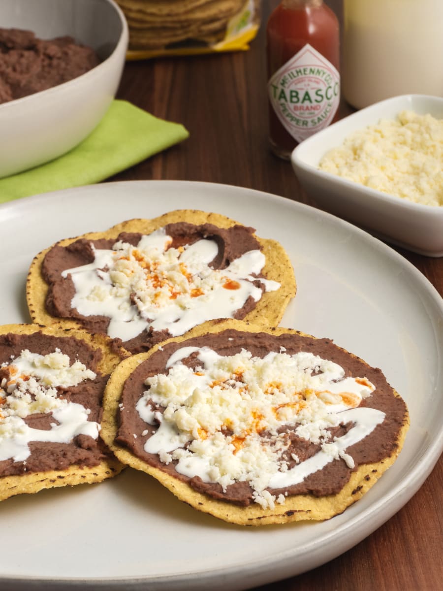 honduran tostadas catrachas served on a plate with toppings like crumbled cheese, tabasco sauce, refried beans, and cream sitting on the counter in the background