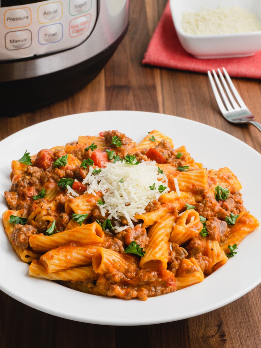 plate of instant pot creamy sausage rigatoni topped with parsley and shredded parmesan cheese with an instant pot in the background a fork and a small bowl of parmesan cheese