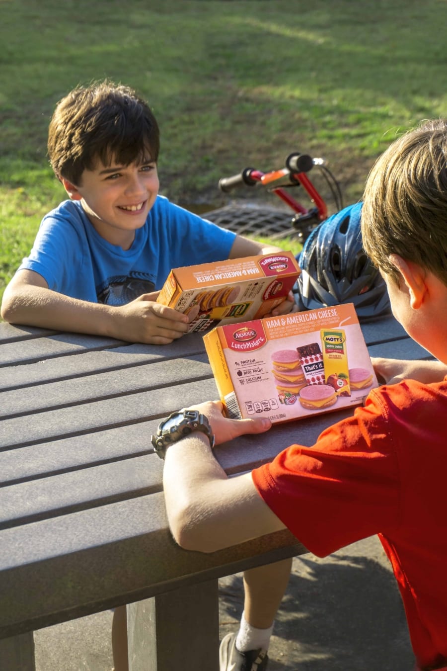 2 kids sitting on park bench each holding a box of armour lunchmakers and drink ham and american cheese