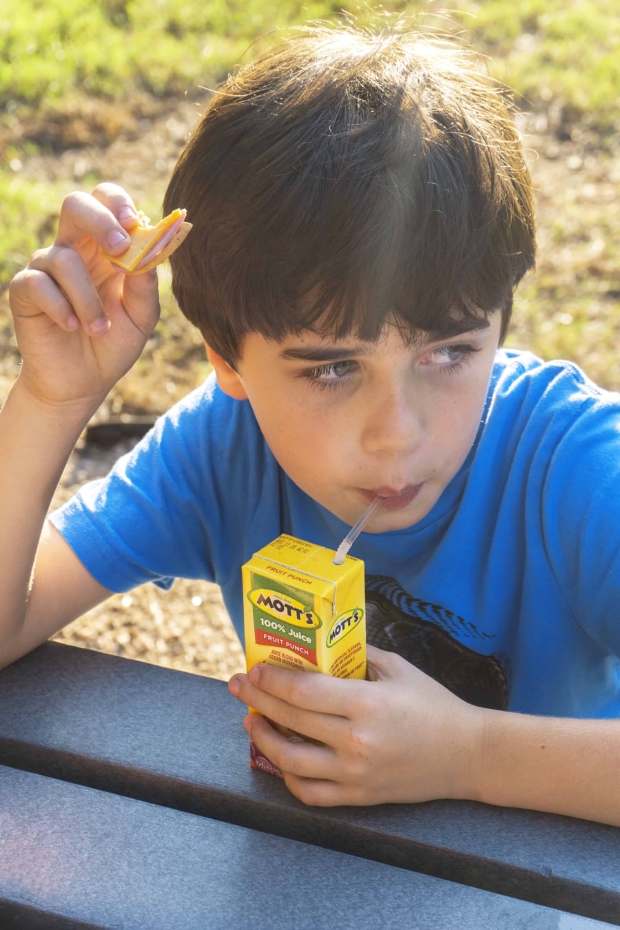 child with blue shirt drinking from a juice box inside and holding a cracker with ham and cheese from a lunchmakers pack