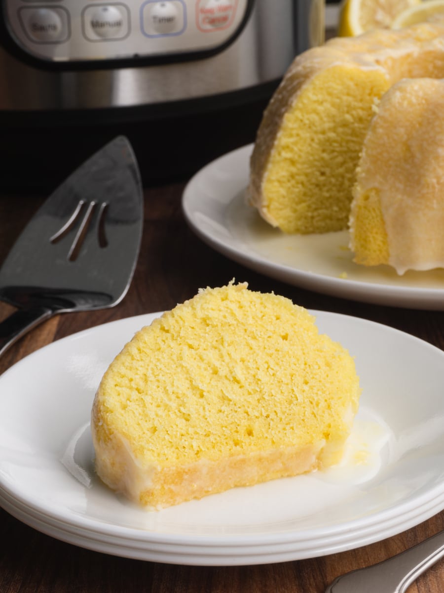 a slice of moist instant pot lemon bundt cake on a white plate with the pressure cooker in the background