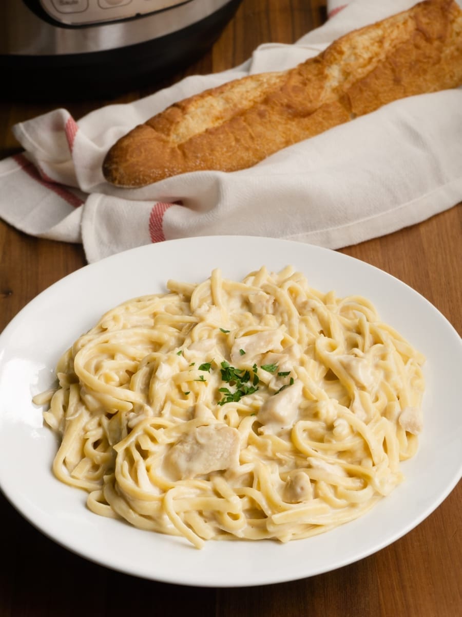 an instant pot in the background with a bowl of creamy chicken fettuccine alfredo up front and some french bread