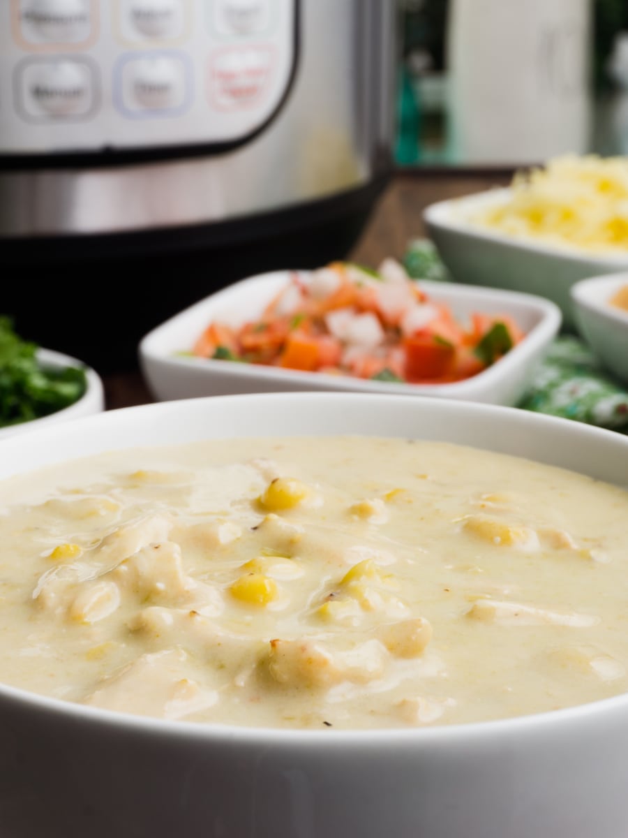 a close up of a bowl of creamy instant pot enchilada soup with corn with the toppings and the instant pot in the background