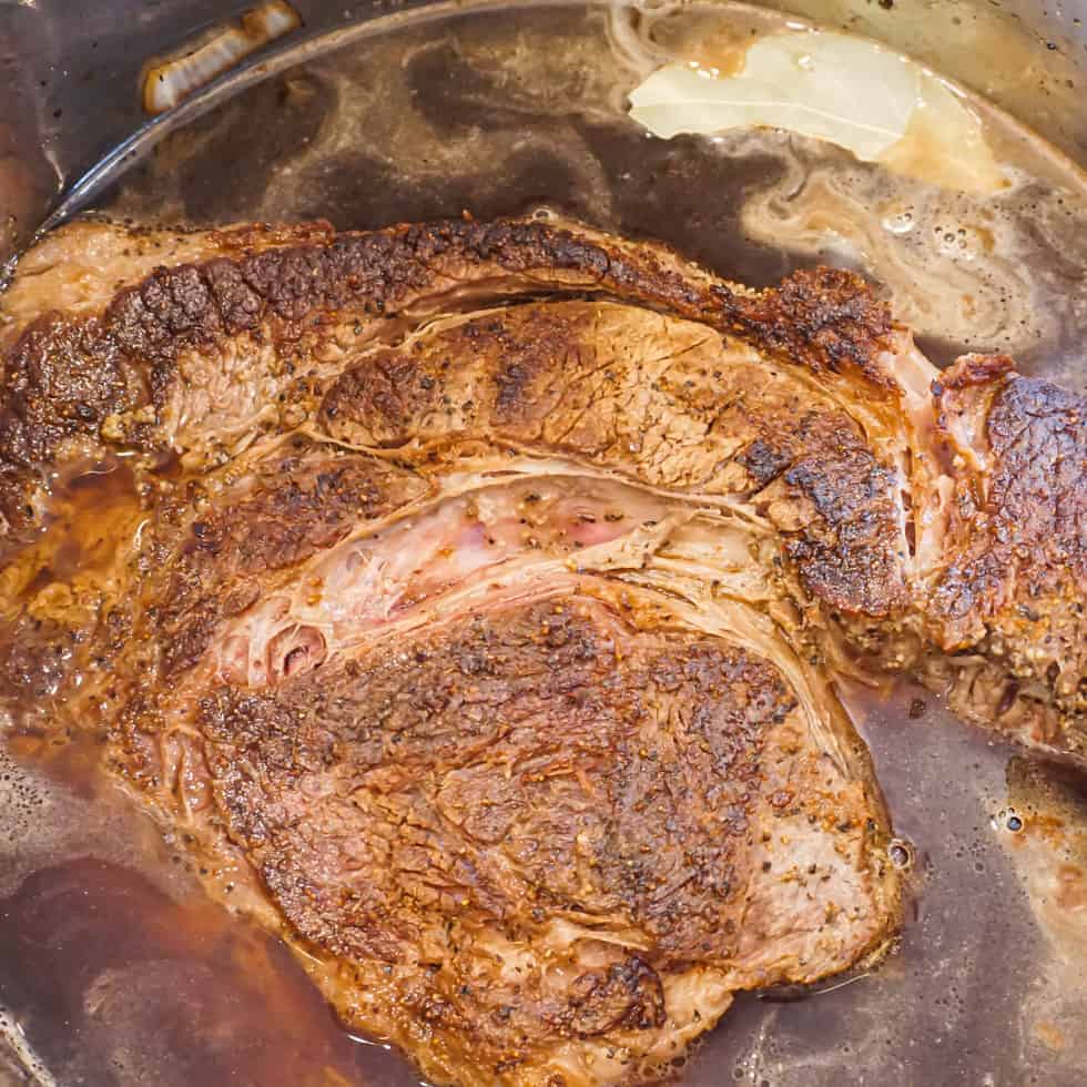 a seasoned and seared chuck roast sitting in a pot of beef broth, red wine, and sliced onions, ready to be pressure cooked