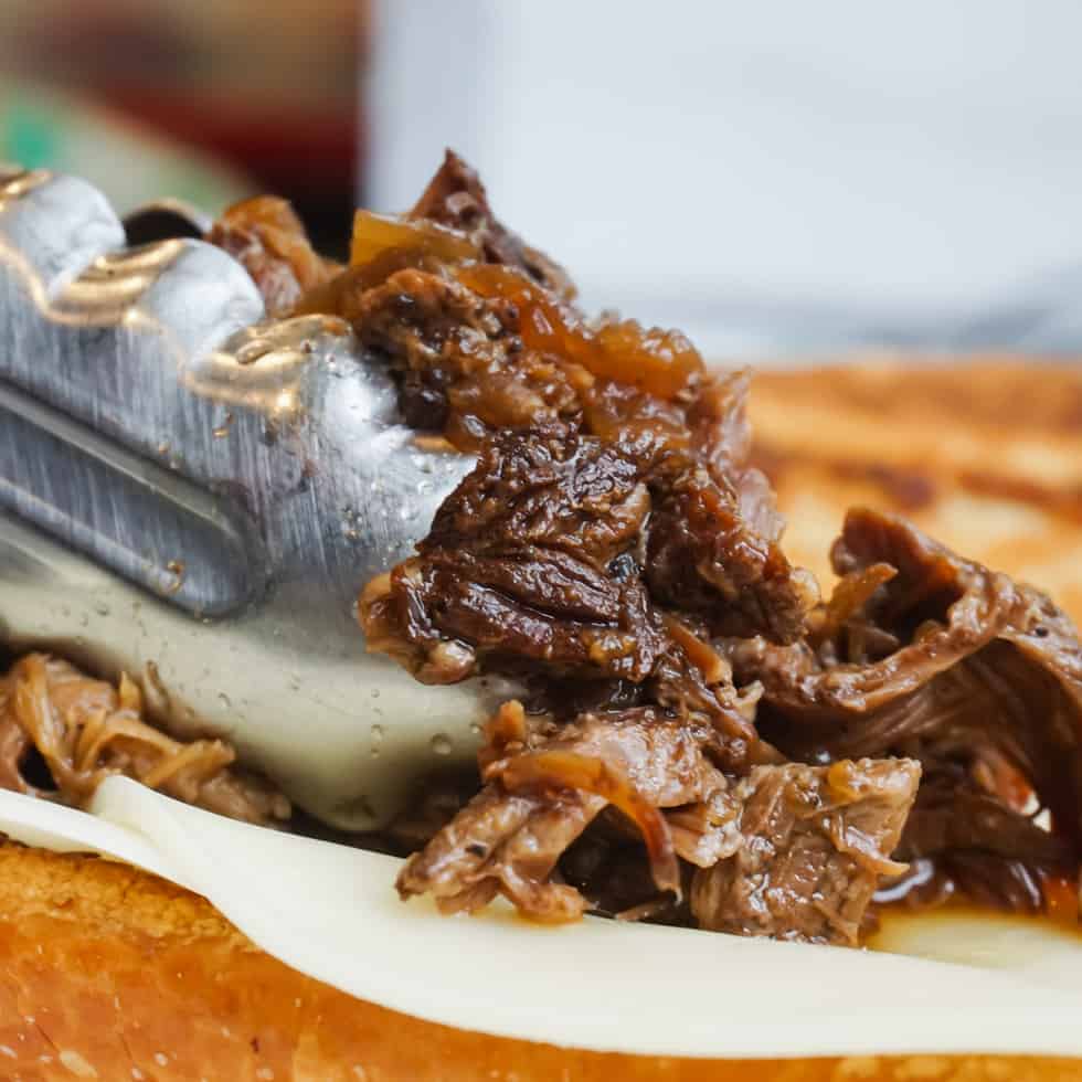 placing cooked chuck roast on a french bread topped with provolone to make french dip sandwiches