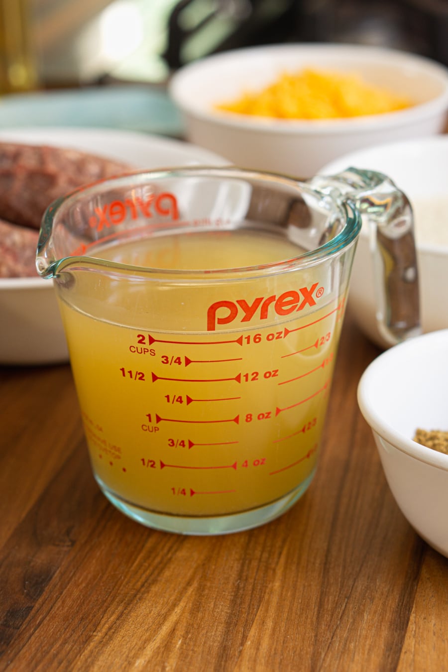 two cups of chicken stock in a measuring cup along with the other ingredients for ground beef and rice casserole including a bowl with two pounds of ground beef, a bowl of shredded sharp cheddar cheese, a bowl of spices, and a bowl of white long-grained rice