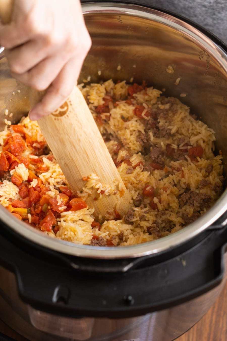 mixing the ingredients of a casserole including ground beef, rice, and rotel using a wooden spatula in the instant pot