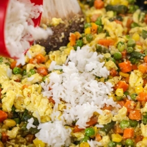 adding a cup of jasmine rice to a skillet filled with mixed vegetables and scrambled egg