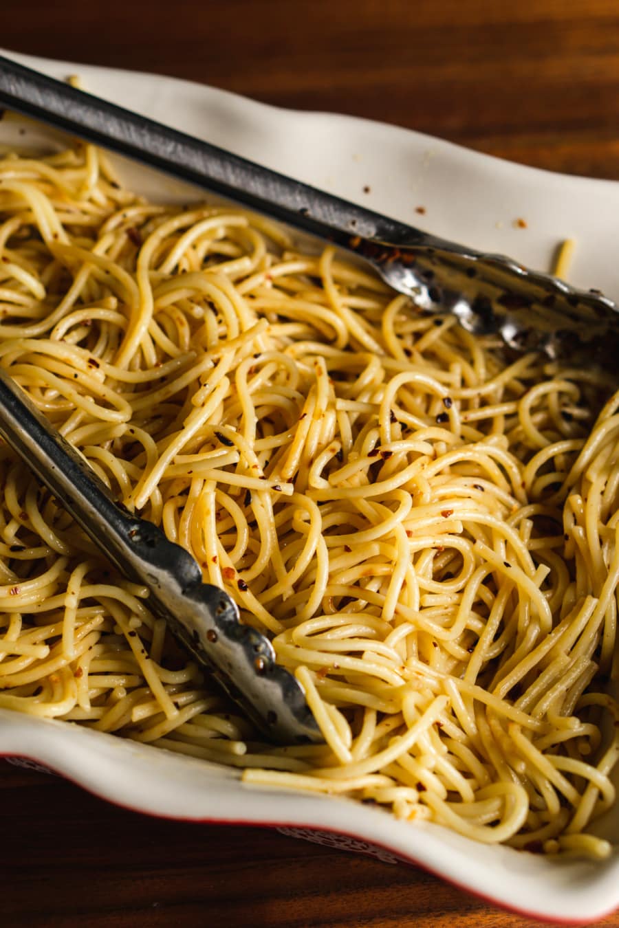 a baking dish filled with spicy pasta aglio e olio with a pair of tongs sitting on top of the pasta