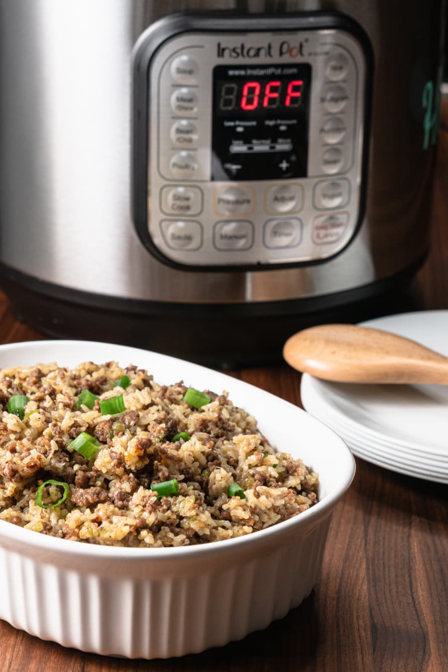 a serving dish with dirty rice, a set of white plates with a wooden spatula and a pressure cooker in the background