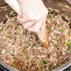 mixing a mixture of cajun seasoned ground beef, ground sausage, and diced vegetables with rice and chicken stock with a large wooden spoon in the instant pot pressure cooker