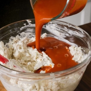 pouring buffalo sauce over a mix of cream cheese, sour cream, mozzarella cheese, shredded chicken, and ranch seasoning in a large glass mixing bowl