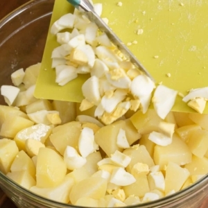 scraping chopped eggs from a cutting board into a bowl of chopped potatoes