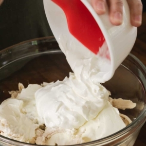 scraping sour cream from a small prep bowl and adding it to a large glass mixing bowl over shredded chicken