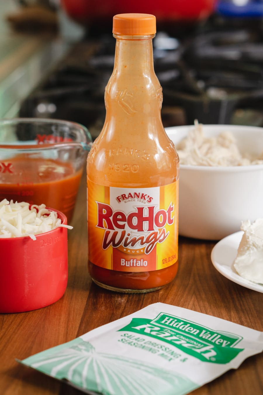 a bottle of Frank's RedHot Wings Buffalo Sauce, a packet of Hidden Valley Ranch salad dressing and seasoning mix, a bowl of shredded chicken, and a cup of shredded mozzarella cheese sitting on the counter