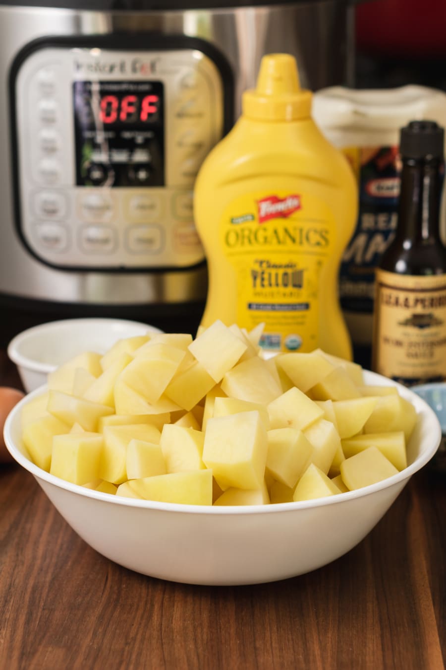 the ingredients for making potato salad sitting on the counter in front of the instant pot including a bowl of chopped potatoes, a bottle of yellow mustard, a bottle of mayonnaise, and a bottle of Worcestershire sauce