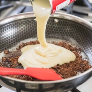 pouring a can of sweetened condensed milk into a warm skillet of melted butter and cocoa powder