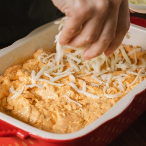 spreading shredded mozzarella cheese over buffalo ranch chicken dip in a casserole