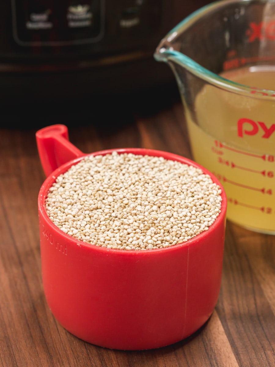 a red plastic measuring cup filled with quinoa with a measuring cup of filled with chicken stock in the background