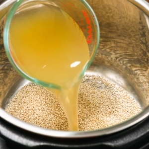 pouring a cup of chicken stock into an electric pressure cooker that has quinoa in it