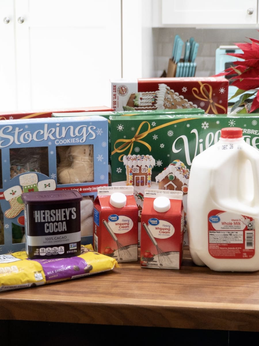 groceries including a gallon of milk, a bag of semi-sweet chocolate morsels, a container of cocoa powder, two small cartons of heavy cream, and four gingerbread house kits sitting on a counter top