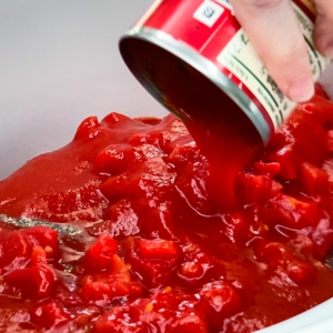 pouring a can of tomato sauce over the ingredients for carne mechada in the crock pot