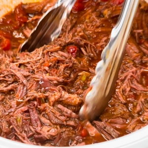 using tongs to grab carne mechada shredded beef from the crockpot