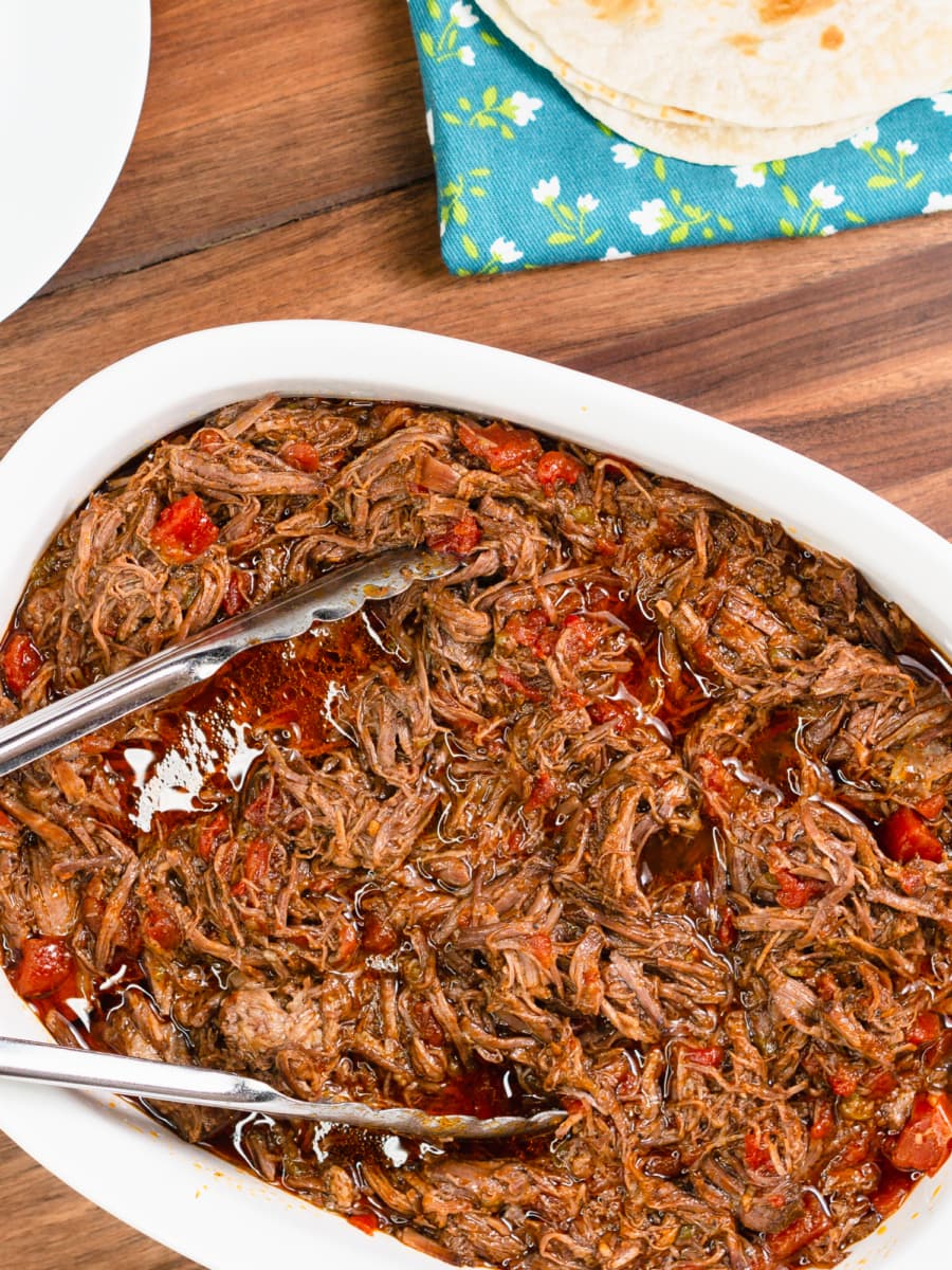 a serving bowl filled with crockpot carne mechada next to a stack of flour tortillas