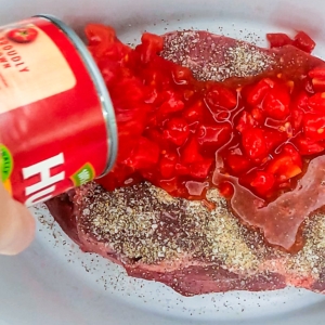 pouring a can of diced tomatoes over a chuck roast in a slow cooker