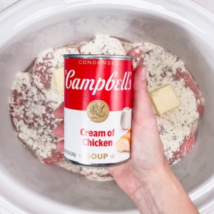 holding a can of cream of chicken over a slow cooker filled with seasoned pork chops