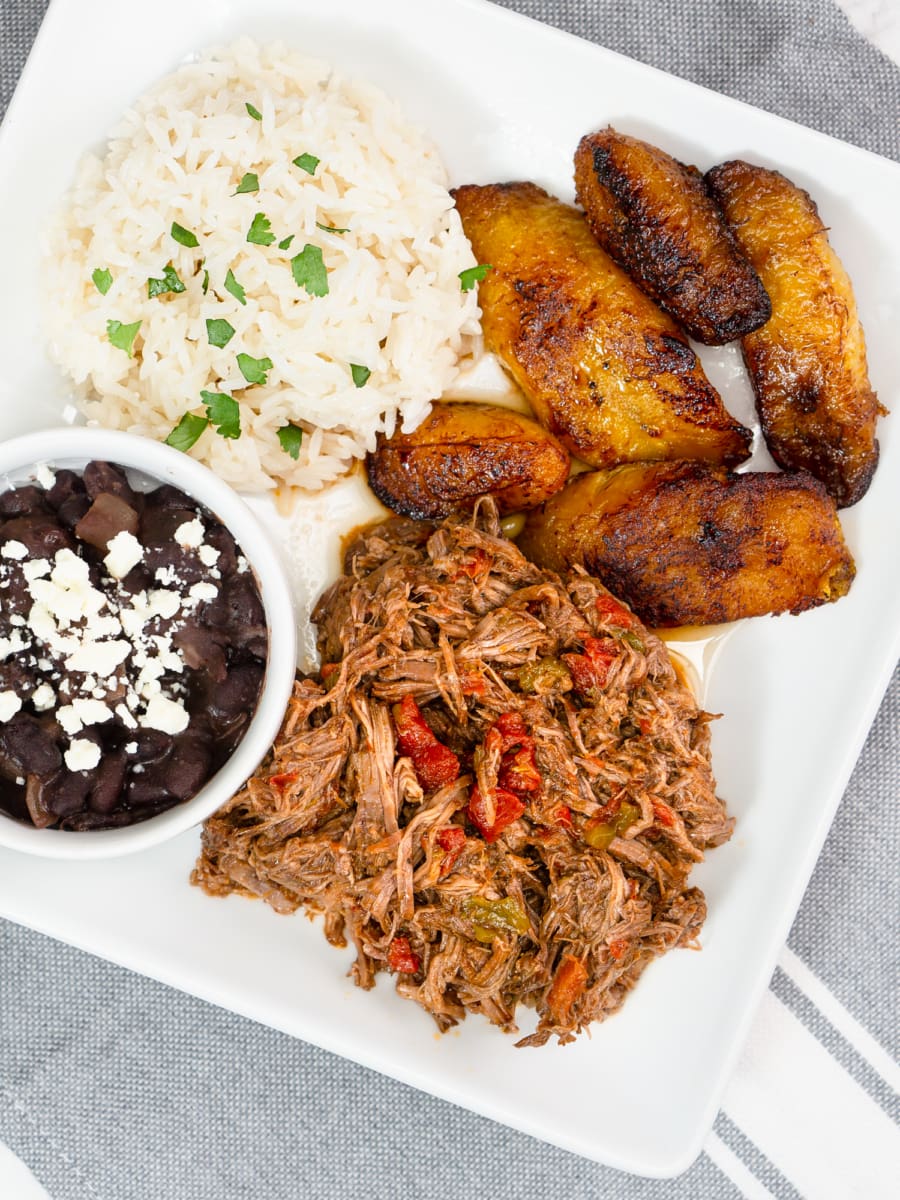 a plate of carne mechada, black beans topped with crumbled white cheese, white rice, and fried plantains